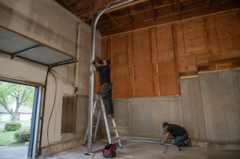 One person on a stepladder working beside a tall curved garage-door track while another kneels beside loose track sections in an unfinished garage.